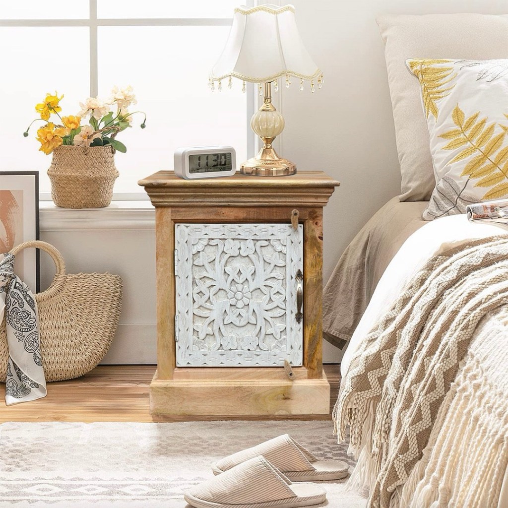 Ręcznie rzeźbiona, bielona szafka nocna z litego drewna mango w stylu boho, ustawiona w jasnej, przytulnej sypialni obok łóżka z beżową pościelą i lampką stołową w klasycznym stylu.
