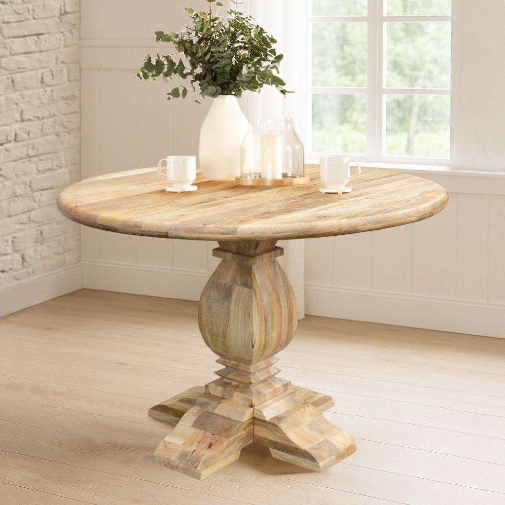 Okrągły stół drewniany Ruth wykonany z litego drewna mango w naturalnym wykończeniu, z ozdobną toczoną podstawą w stylu indyjskim, ustawiony w jasnej jadalni z dekoracjami.