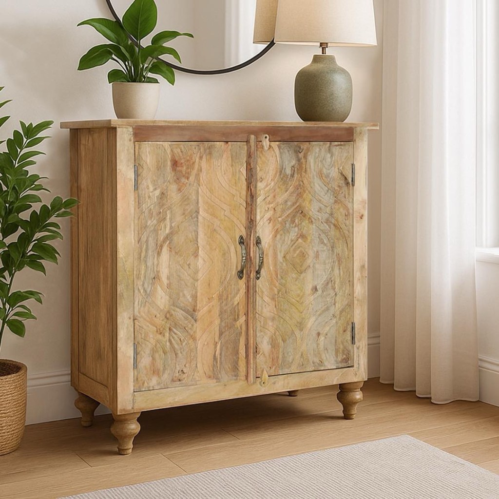 Mała komoda indyjska wykonana z jasnego drewna mango. Fronty dwudrzwiowej szafki są ręcznie rzeźbione w delikatny, geometryczny wzór orientalny. Mebel stoi w jasnym, minimalistycznym przedpokoju.